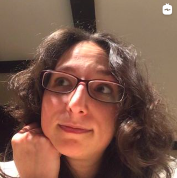 Erica with brown wavy frizzy hear and burgundy glasses resting her head on her right hand looking up to her right curiously with a slight smile on her face.