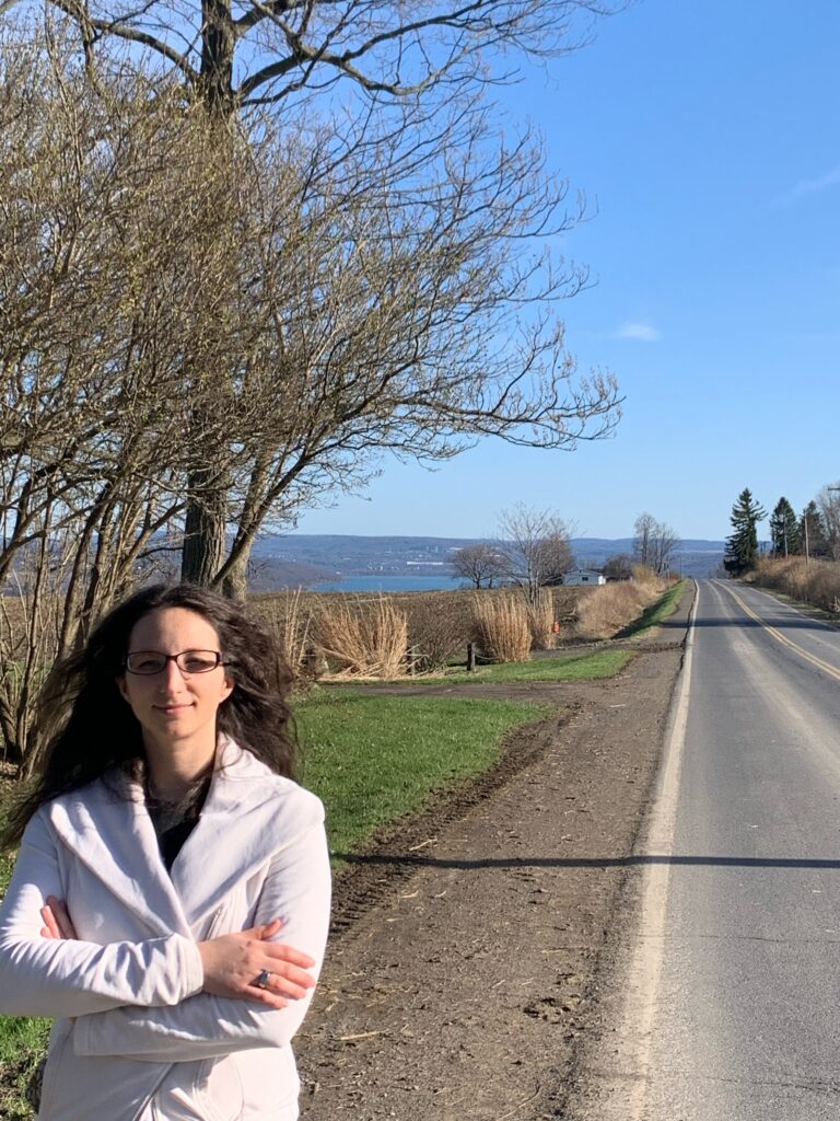 Erica with long brown hair stands at the edge of a rural road, arms crossed, looking toward the camera. A bare-branched tree is behind her and a lake view stretches in the distance. Early spring.