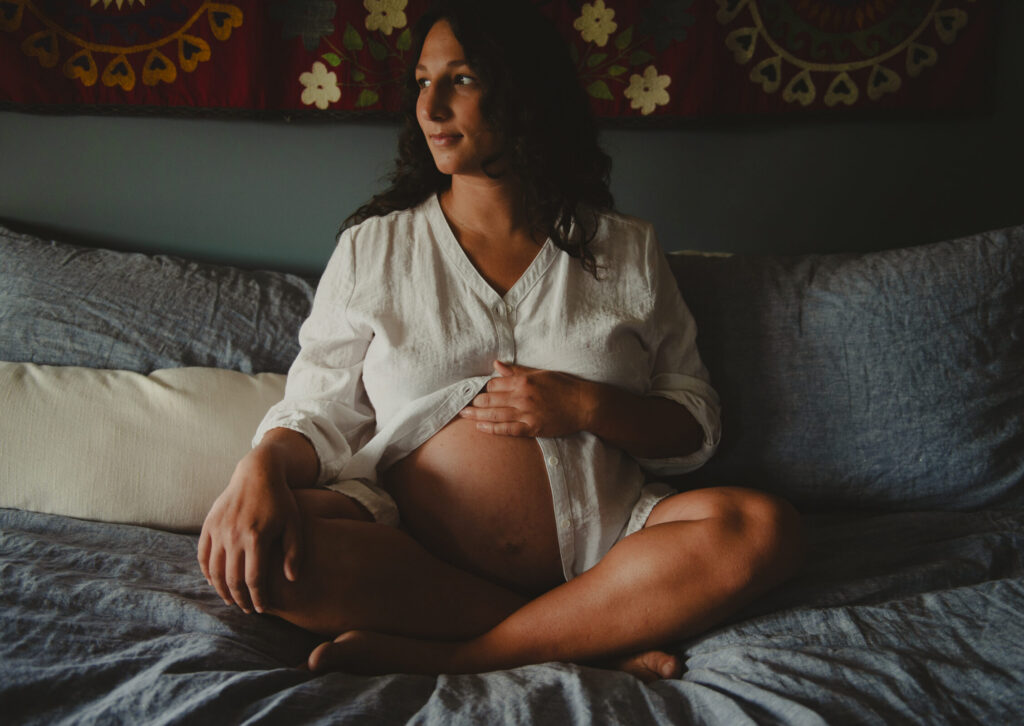 Erica Evans sitting cross-legged on a bed during pregnancy, looking thoughtfully out a nearby window while resting a hand on her belly.